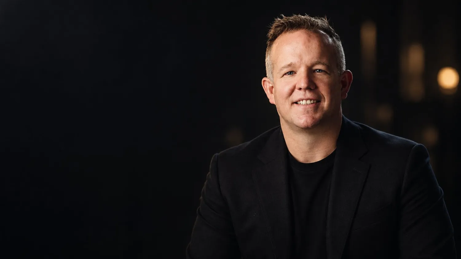 Jon Morrison, seated in a dark studio, smiling toward the camera in a black jacket